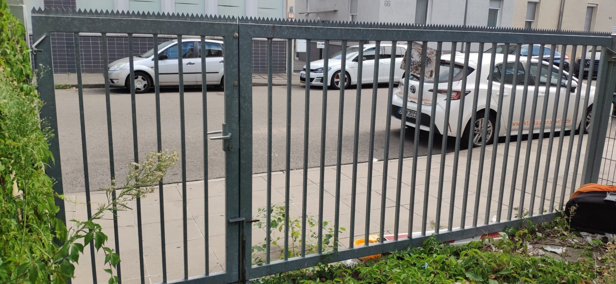 Metallzaun mit senkrechten Streben und einer Öffnung, im Hintergrund parkende Autos auf einer Straße.