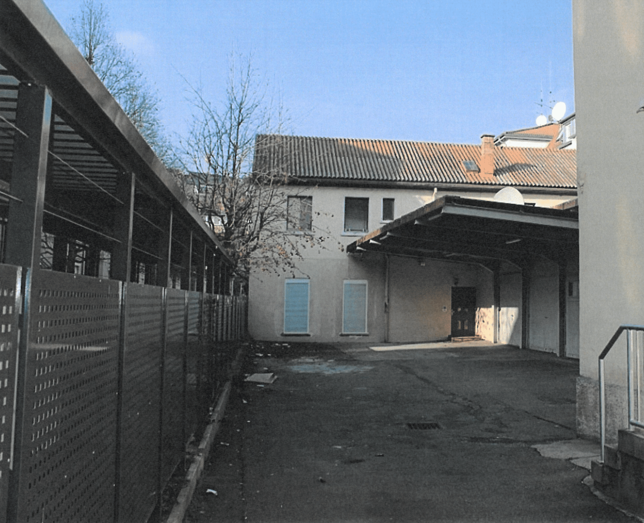 Hinteransicht eines Gebäudes mit betoniertem Hof und wenigen Bäumen, im Hintergrund ein blauer Himmel.