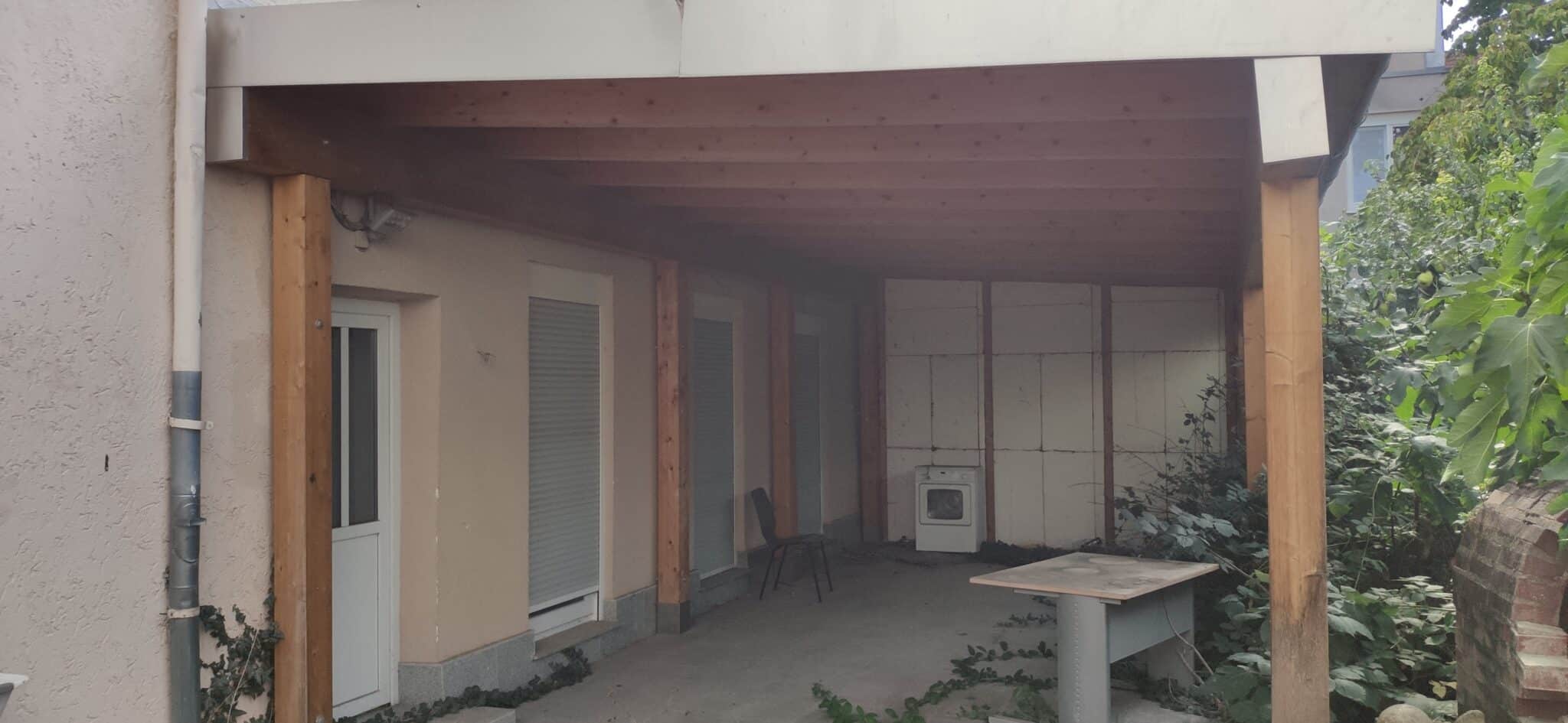 Überdachte Terrasse mit Holzstützen, neben einer Wand und umgeben von Pflanzen, ohne Möbel außer einem Tisch und einem Stuhl.