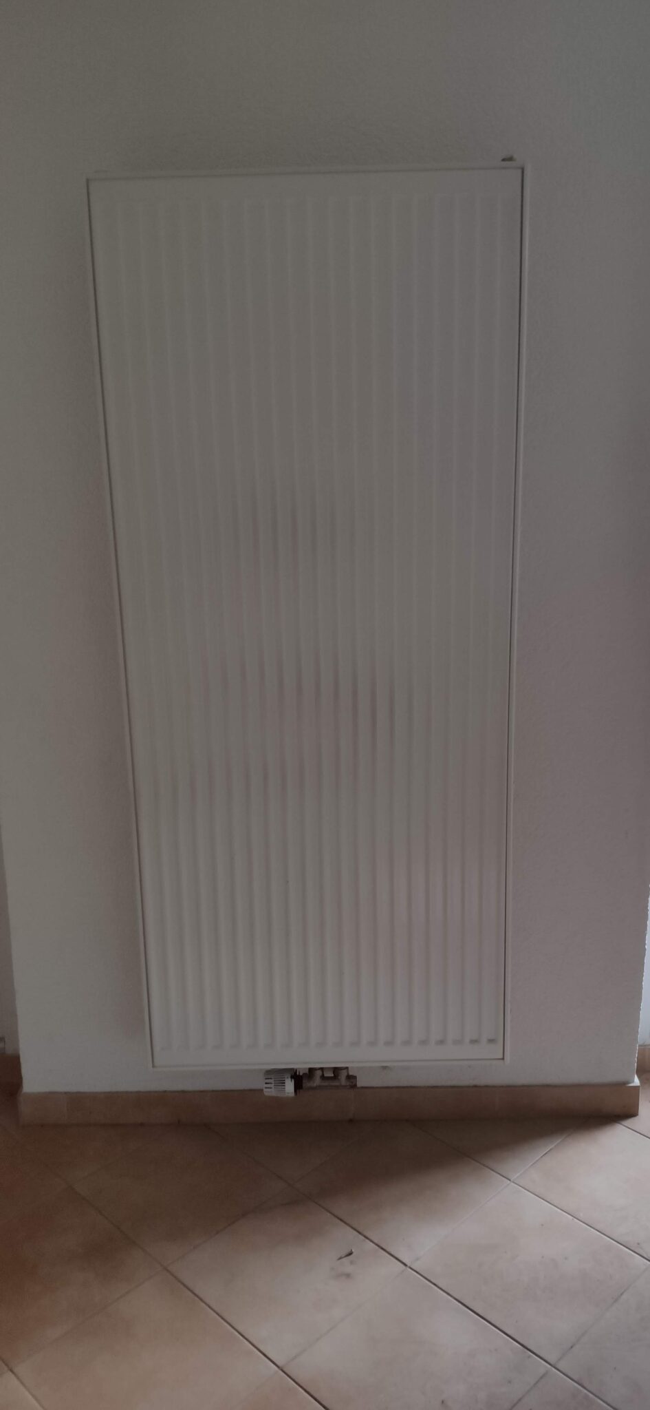 Ein vertikaler Heizkörper an einer weißen Wand, vor einem hellen Fliesenboden.