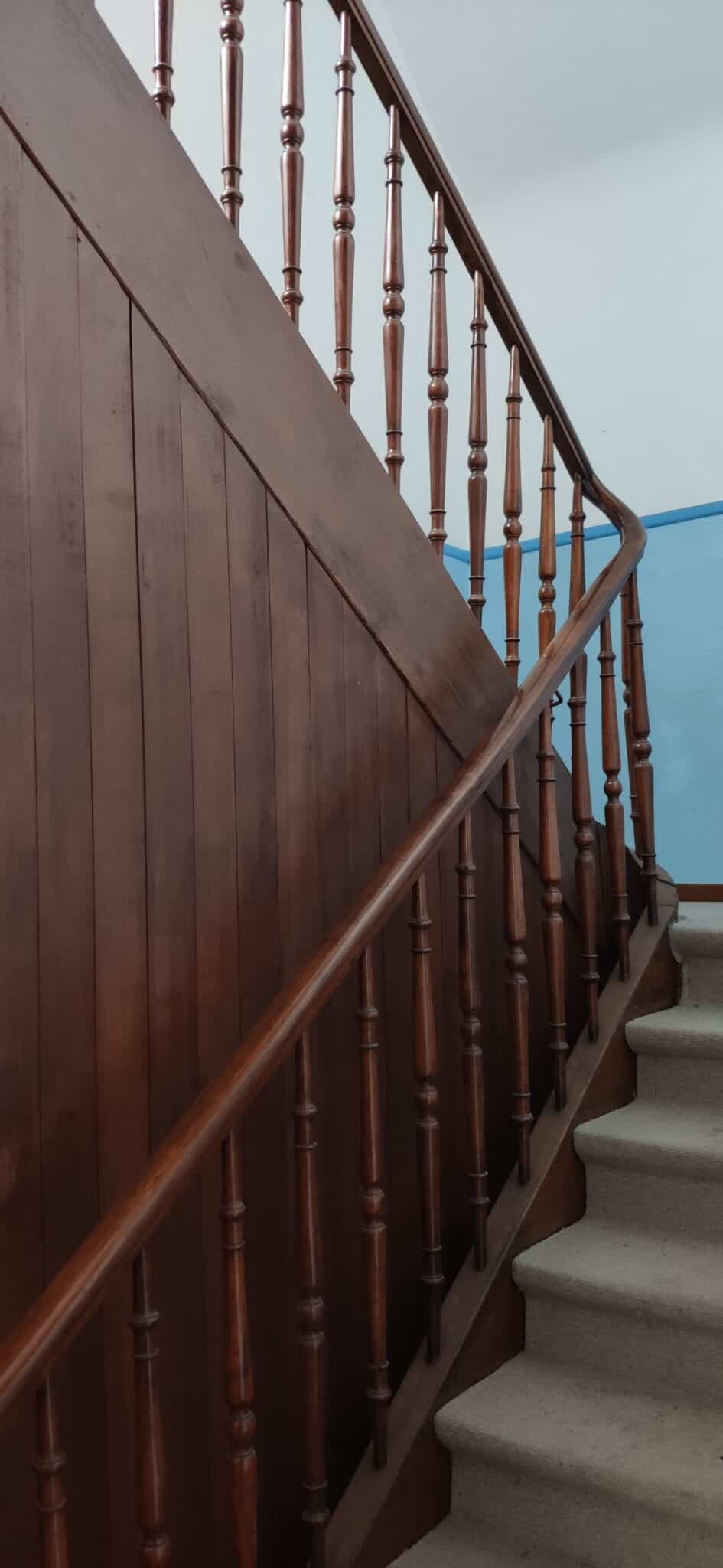 Holztreppe mit balustradierten Geländer und blau gestrichener Wand im Hintergrund.