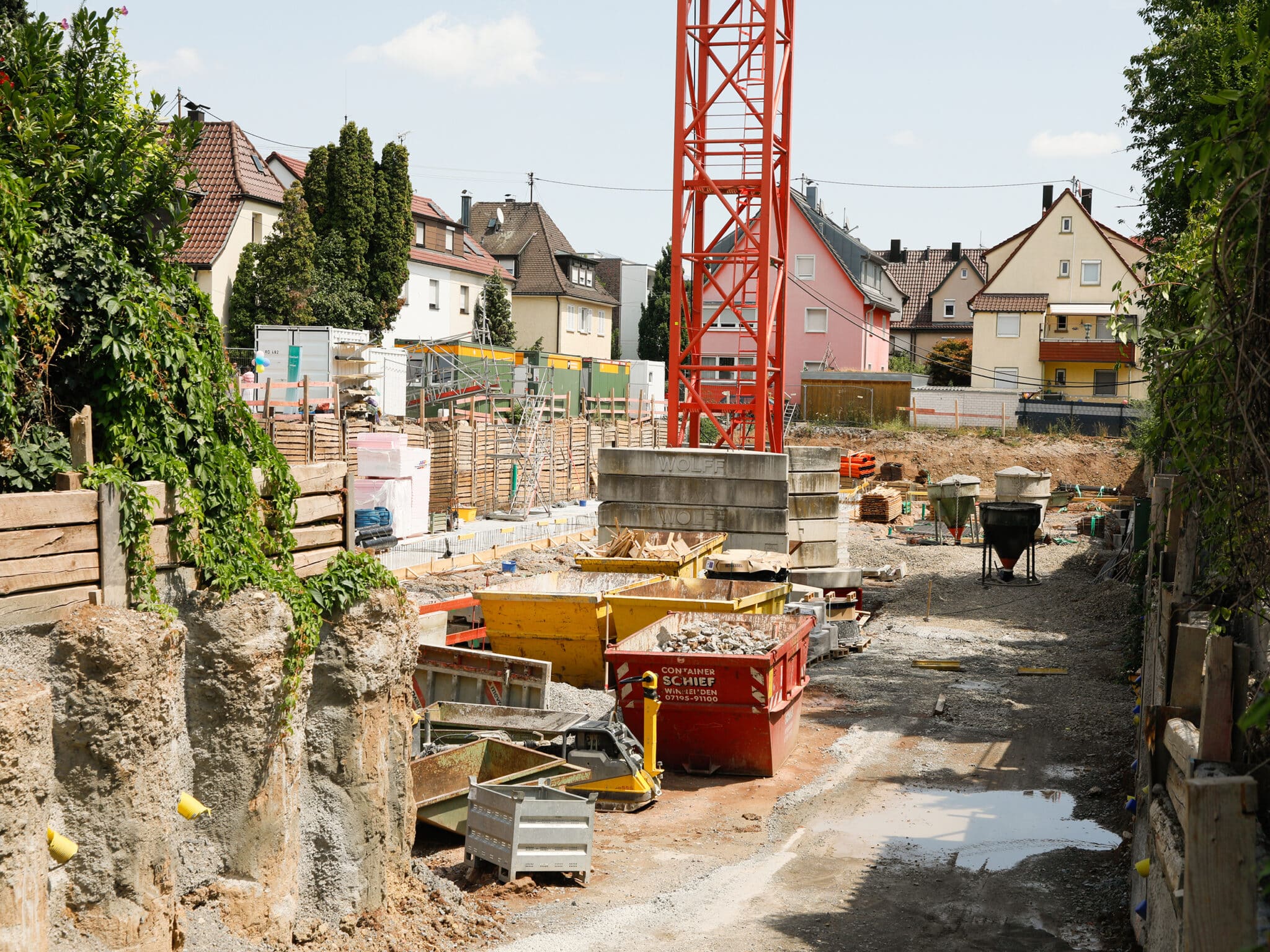Eine Baustelle mit Baugrube und rotem Baukran erstreckt sich zwischen bestehenden Wohnhäusern. Im Vordergrund sind Container, Baumaterialien und Arbeitsgeräte zu sehen, während Schalungen und Fundamente den Baufortschritt erkennen lassen.