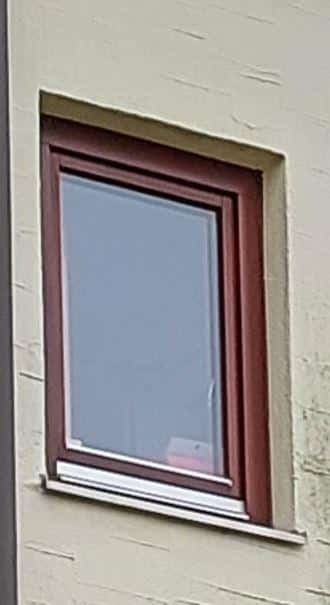 Ein braunes Fenster in einer beige gestrichenen Wand, leicht geöffnet mit Sicht auf einen grauen Innenraum.