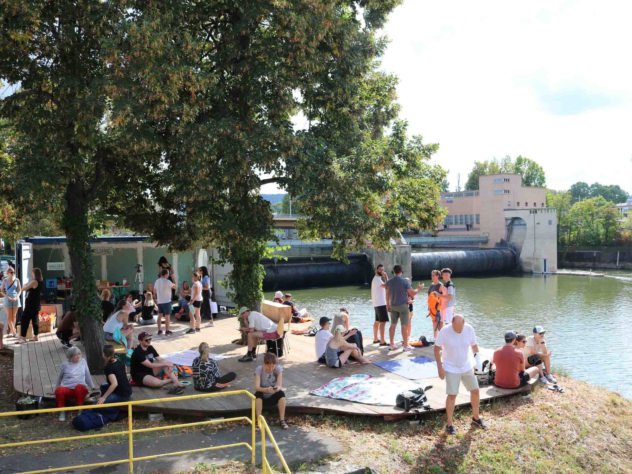 Menschen sitzen auf einem Sonnendeck unter einem Baum mit Blick auf den Neckar und das Stauwehr.