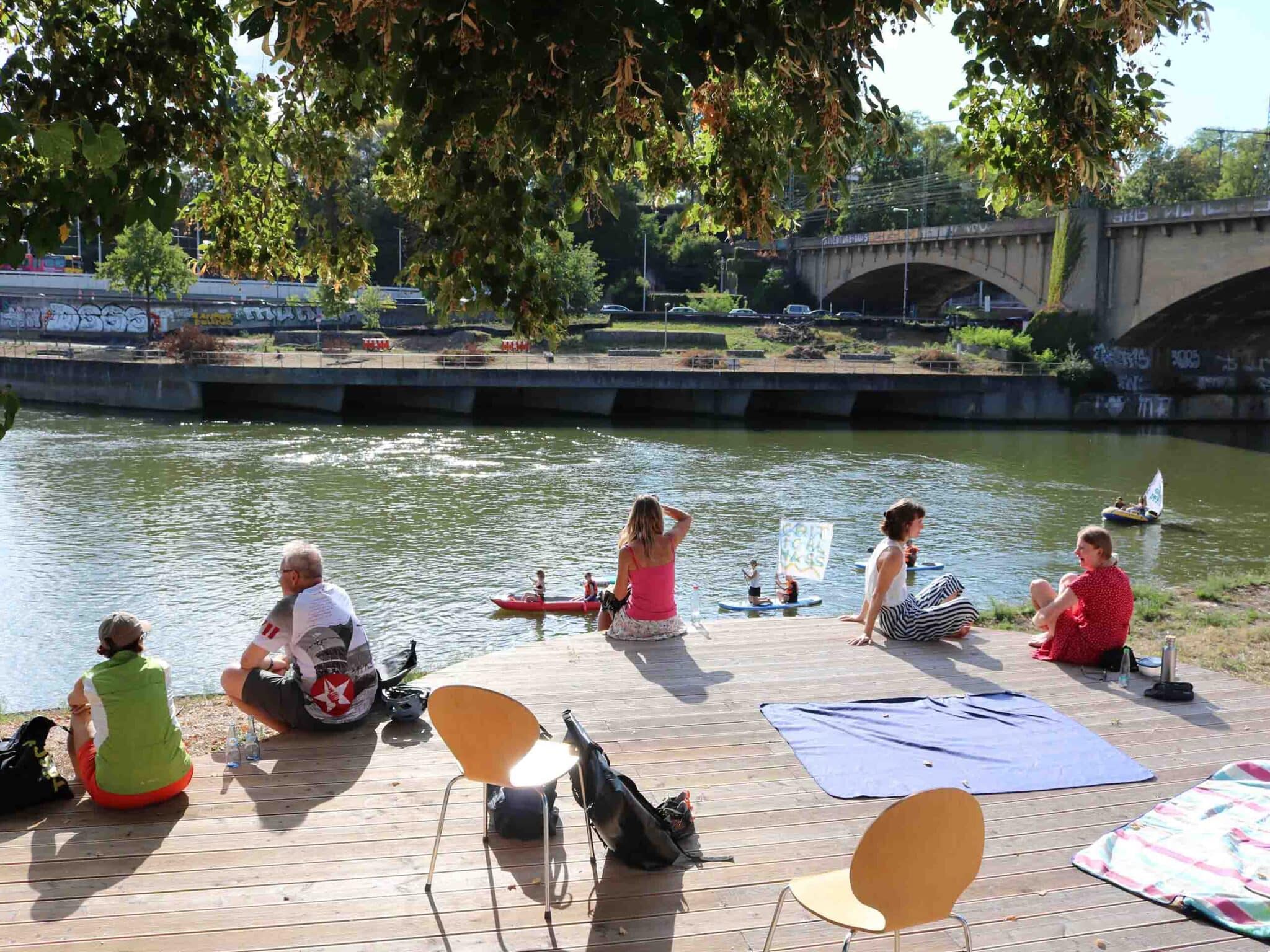 Menschen sitzen bei Sonne auf einem Holzdeck und schauen auf den Neckar.