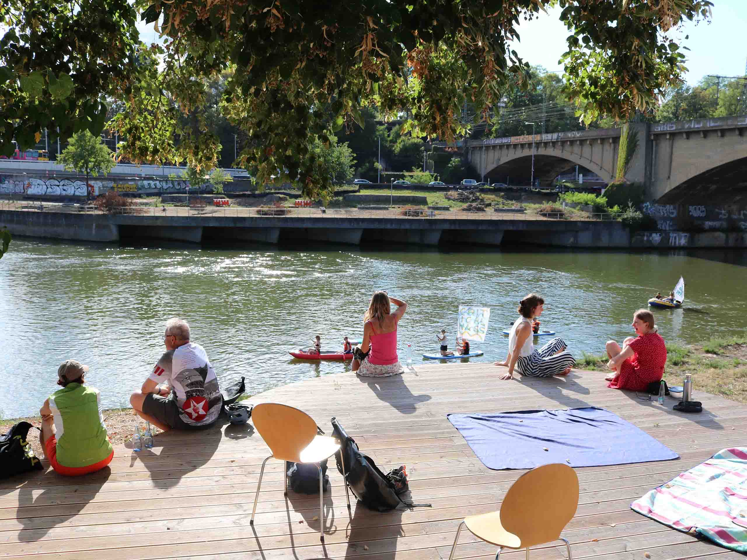 Menschen sitzen bei Sonne auf einem Holzdeck und schauen auf den Neckar.