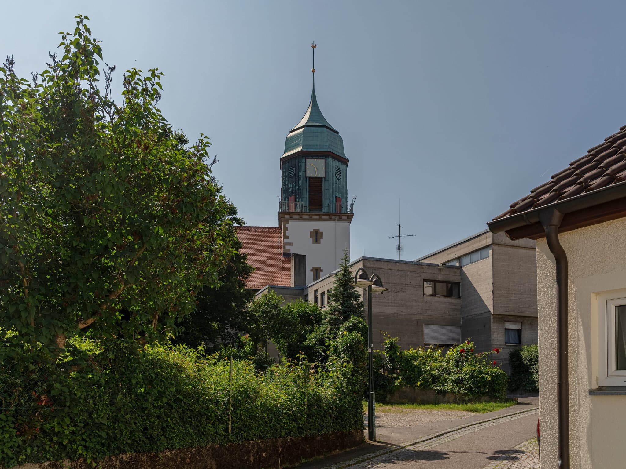Turm mit grüner Kuppel, umgeben von Bäumen und modernen Gebäuden, unter blauem Himmel.