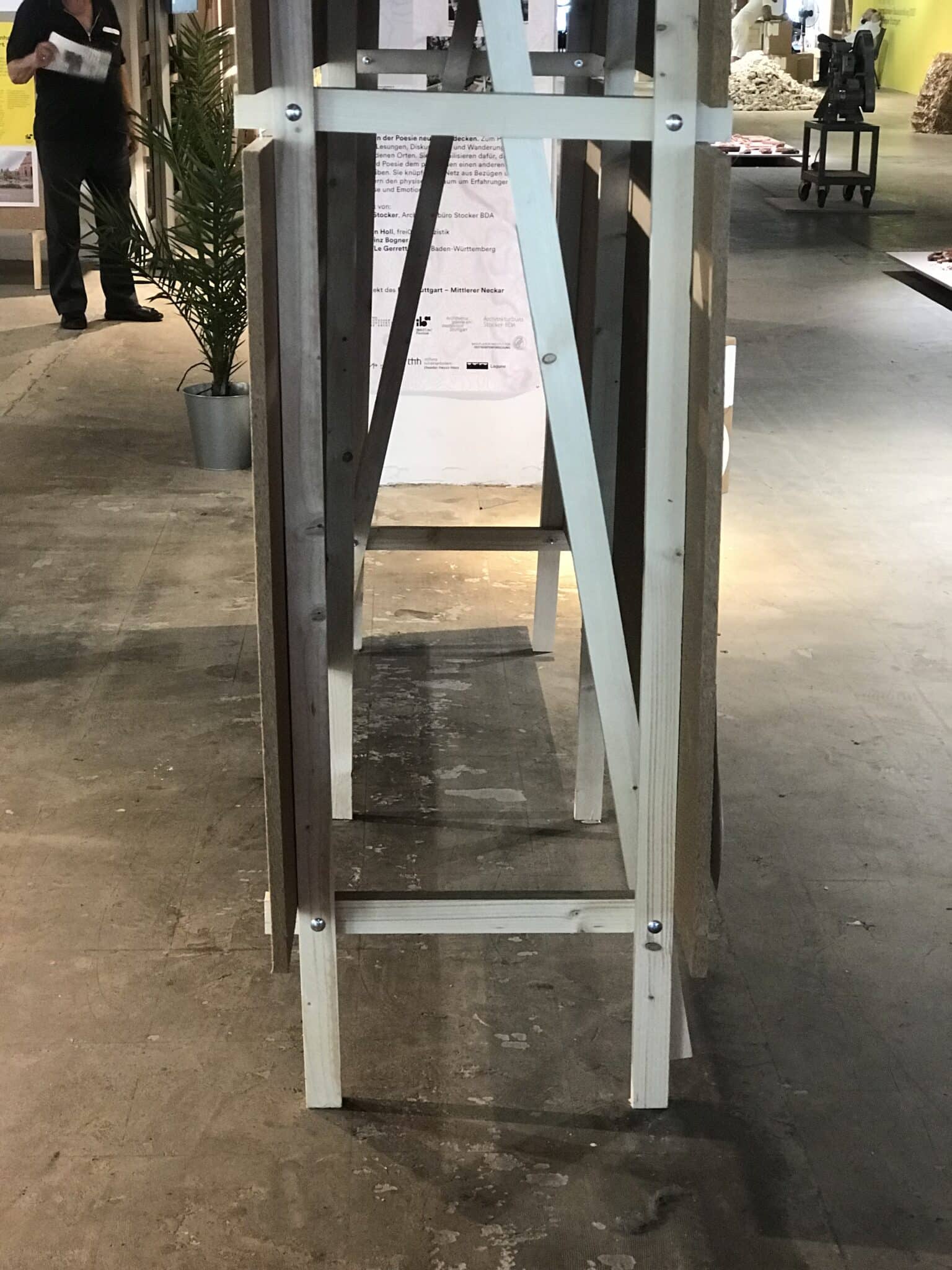 Holzgestell in einem Ausstellungsraum, links im Bild ist eine Pflanze in einem Topf zu sehen.