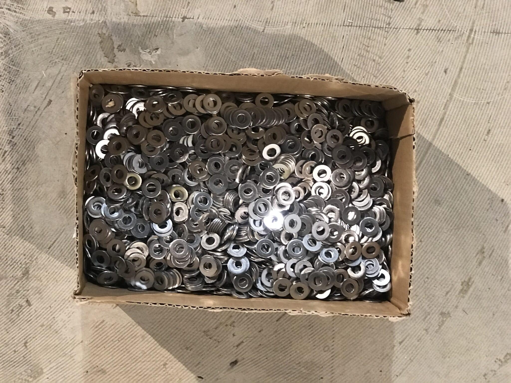 Eine Kiste voller runder Metallunterlegscheiben auf einem grauen Untergrund.