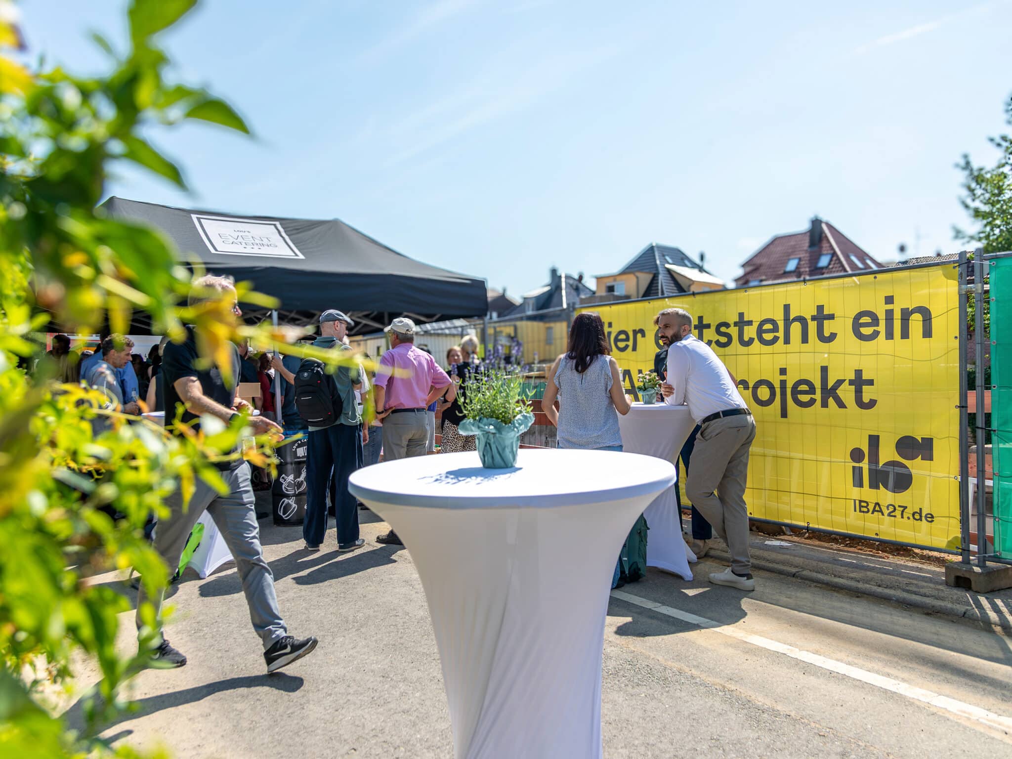 Mehrere Gäste stehen an Stehtischen vor einer Baustelle und unterhalten sich bei einer Veranstaltung im Freien. Rechts verläuft ein Bauzaun mit einem großen gelben Banner, links bietet ein Pavillon Schatten. Im Vordergrund steht ein mit einer kleinen Blumendekoration geschmückter Stehtisch, dahinter sind Wohnhäuser und ein sonniger Himmel zu sehen.