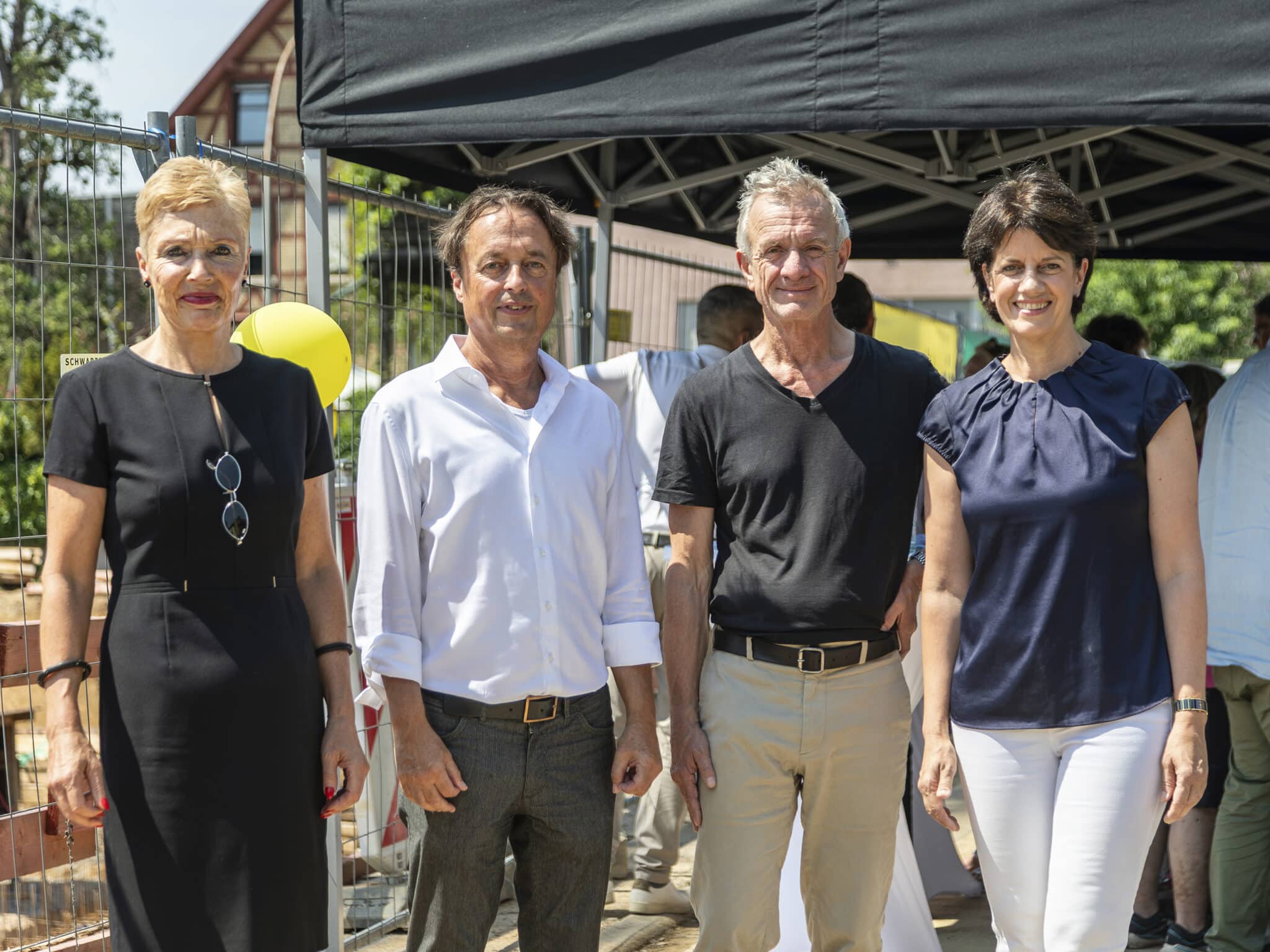 Vier Personen stehen nebeneinander im Freien vor einem schwarzen Pavillonzelt auf einer Baustelle, im Hintergrund ein Bauzaun. Von links: Fellbacher Baubürgermeisterin Beatrice Soltys in schwarzem Kleid, Christoph Welz, Geschäftsführer der Siedlungswerk GmbH, in weißem Hemd, IBA'27-Intendant Andreas Hofer in dunklem T-Shirt und beiger Hose sowie Fellbachs Oberbürgermeisterin Gabriele Zull in dunkelblauer Bluse. Aufnahme vom Baustellenfest Eppinger Straße.