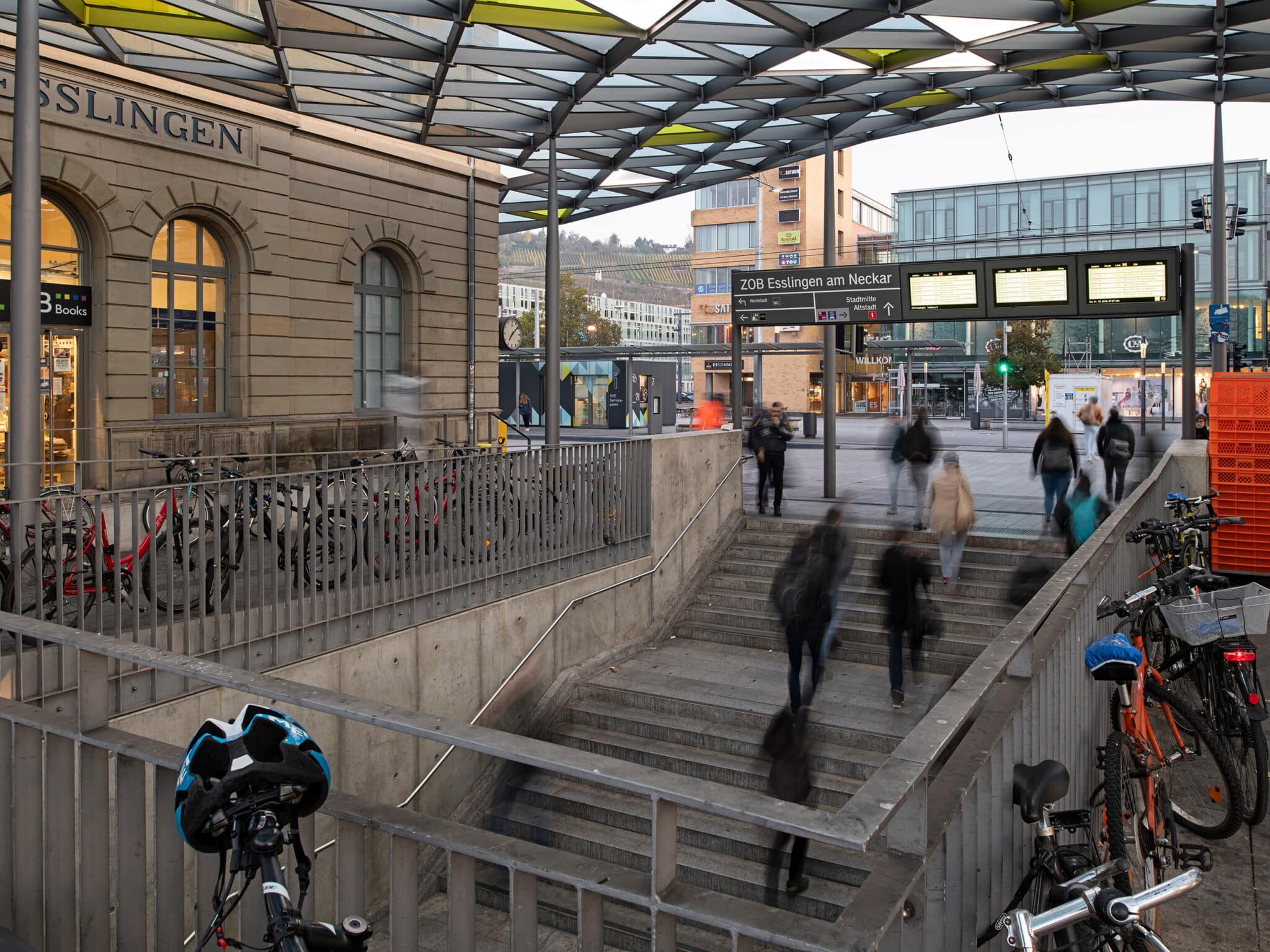 Menschen laufen eine Treppe hinauf und hinunter. Man erkennt eine Bahnhofssituation. An der Seite ist ein alter Gebäude mit Fenstern zu erkennen. Rechts parken Fahrräder.