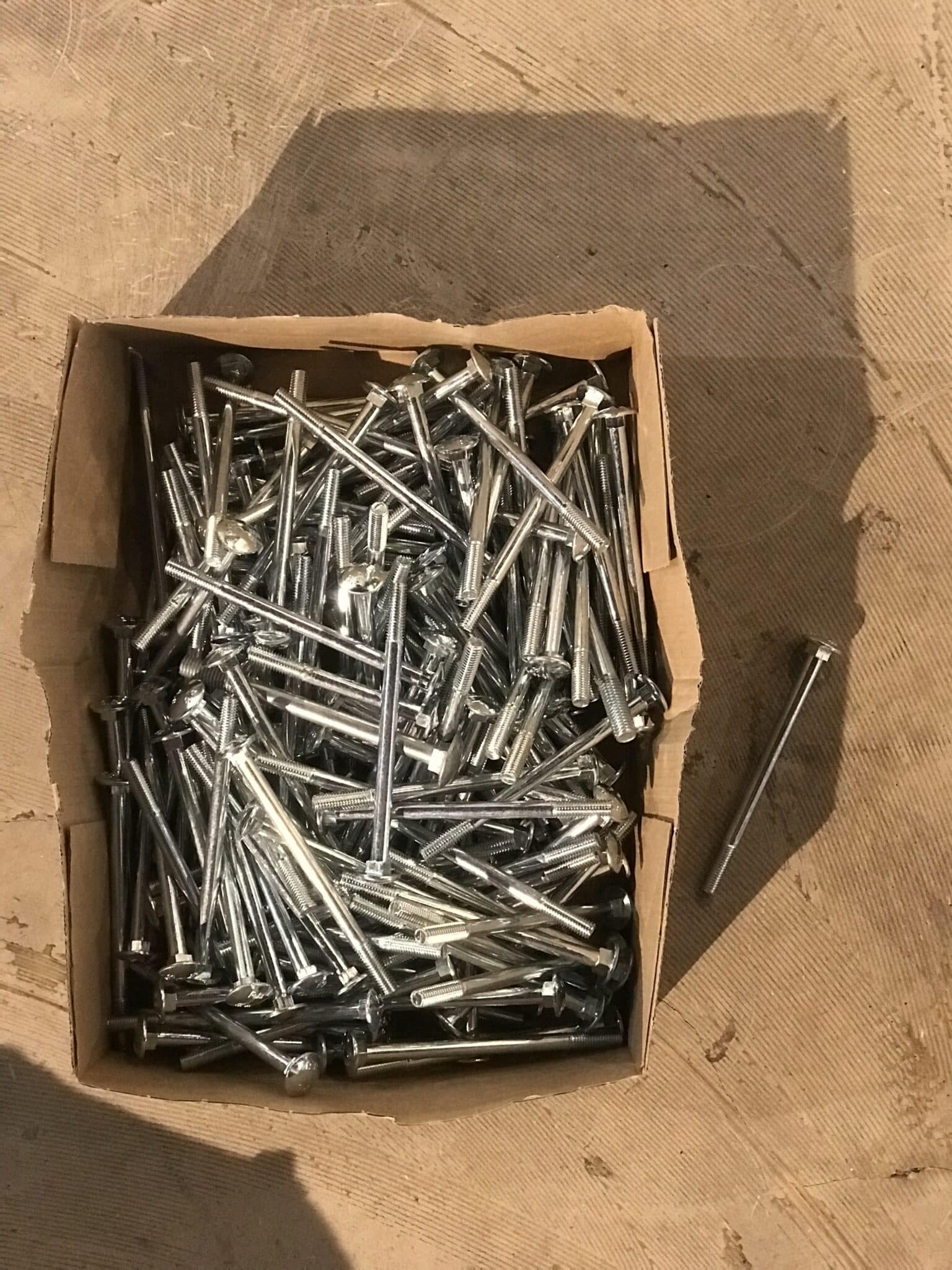 Eine offene Kartonbox gefüllt mit vielen silbernen Schrauben, eine Schraube liegt daneben auf dem Boden.
