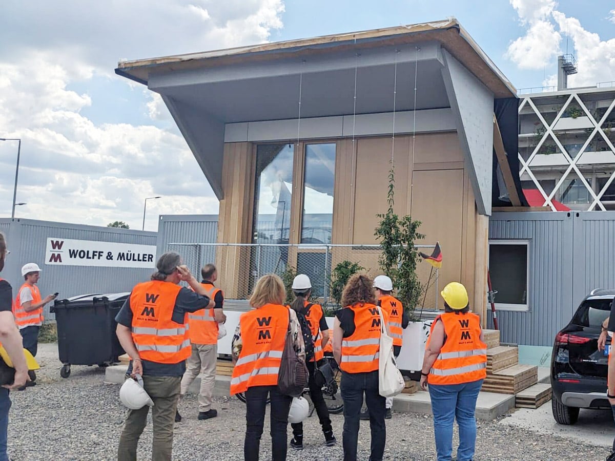 Gruppe von Personen mit Sicherheitswesten und Helmen vor einem modernen Gebäude auf einer Baustelle.