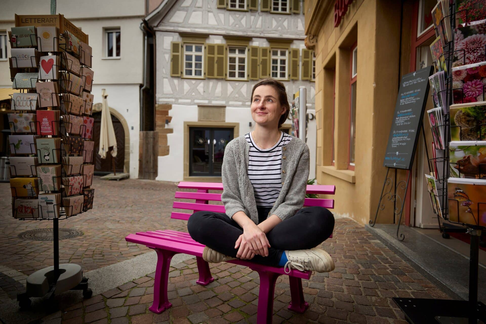 Eine Frau auf einer pinken Bank vor alten Häusern.