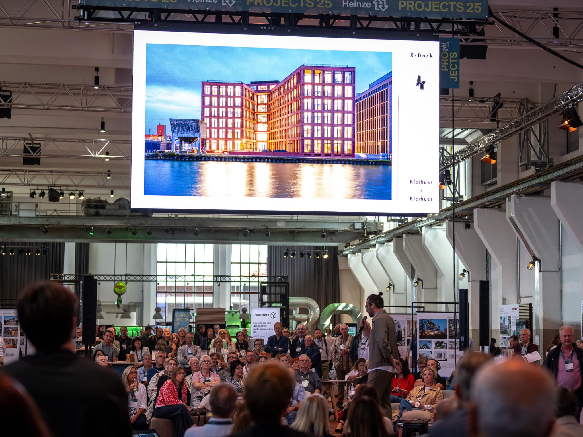 Raum voller Menschen, ein großes Power-Point-Bild von einem beleuchteten Haus zur blauen Stunde.