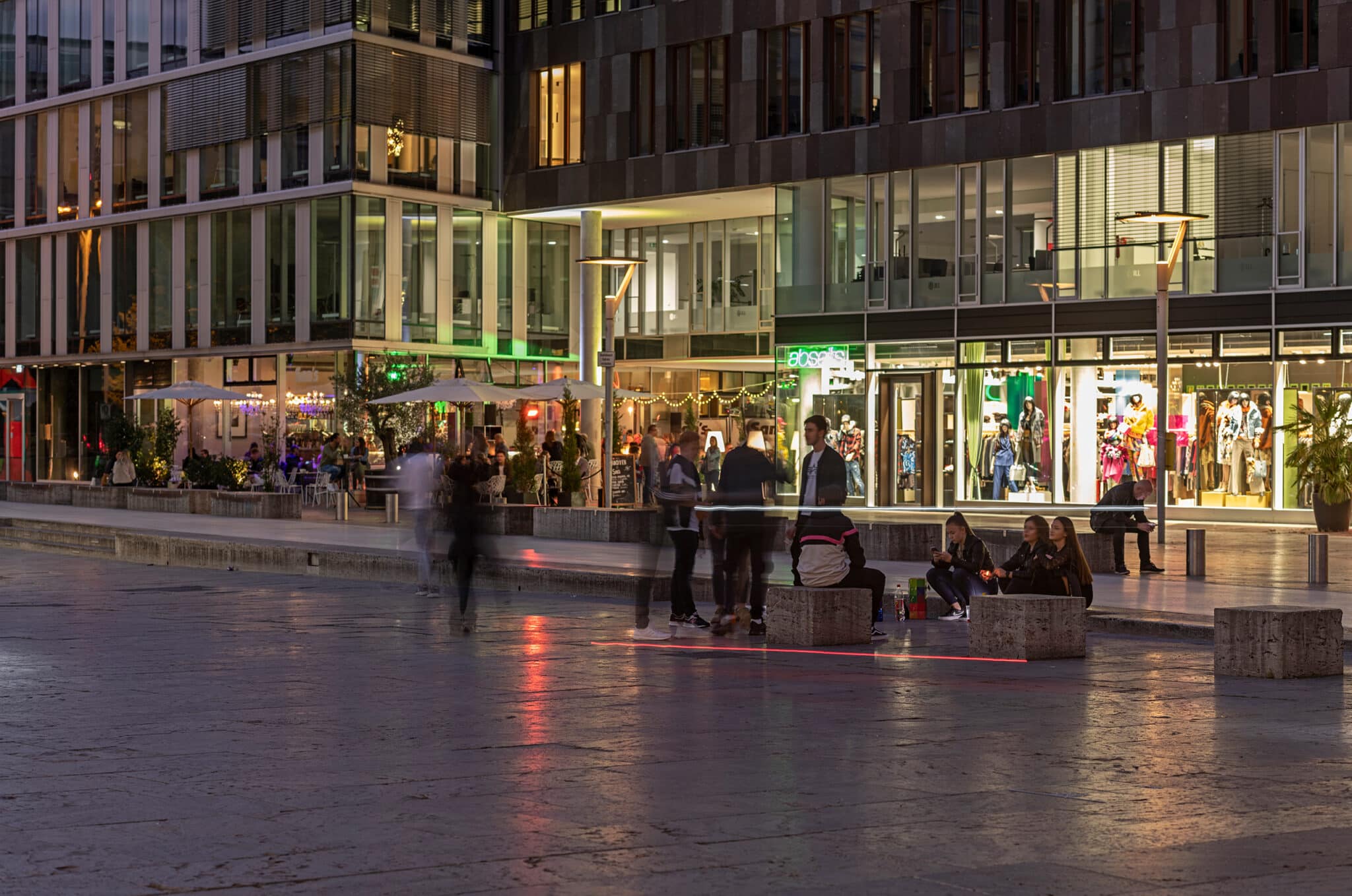 Abendliche Stadtansicht mit Menschen an einem Platz und modernen Gebäuden, beleuchtet von Schaufenstern und Straßenlampen.